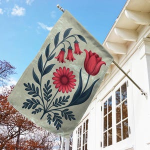 May include: A decorative outdoor flag featuring a floral design. The flag displays a red tulip, daisy, and bell-shaped flowers with dark blue leaves and stems against a light beige background. The flag is hanging outside a white house.