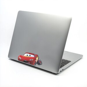 May include: A silver laptop with a red Lightning McQueen sticker on the lower left corner. The sticker features the animated race car with blue eyes and yellow lightning bolt details. The laptop is on a white background.