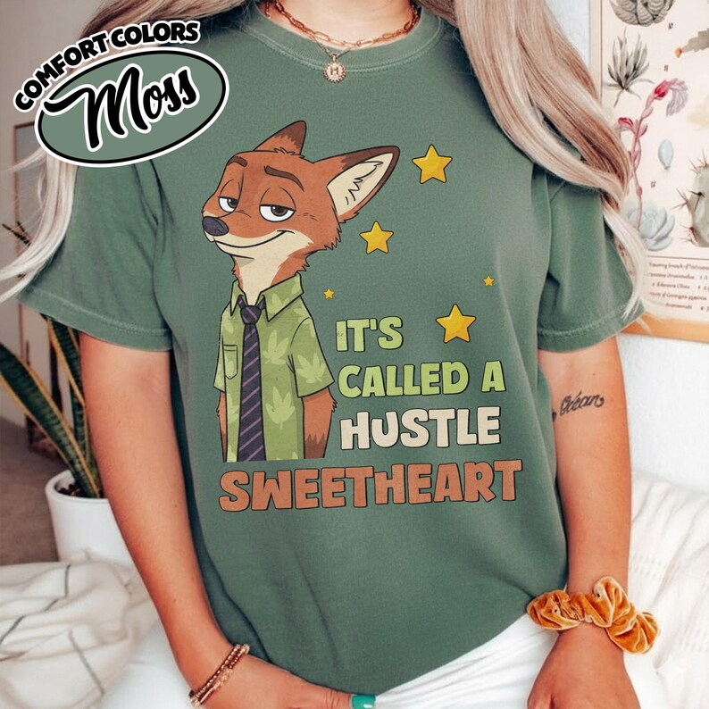 Puede incluir: Una camiseta de manga corta color verde musgo con el gr&aacute;fico de un zorro de dibujos animados que viste una camisa verde y corbata oscura. El texto dice IT'S CALLED A HUSTLE SWEETHEART, junto a estrellas amarillas. Tiene cuello redondo.