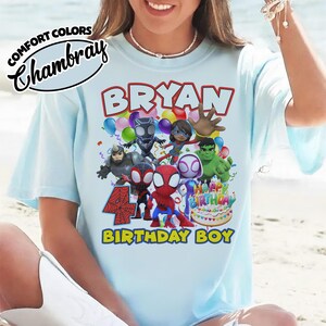 May include: A light blue t-shirt featuring a graphic print with the text BRYAN, 4, and BIRTHDAY BOY. The design includes cartoon superhero characters, colorful balloons, and a birthday cake with the text HAPPY BIRTHDAY. COMFORT COLORS CHAMBRAY.