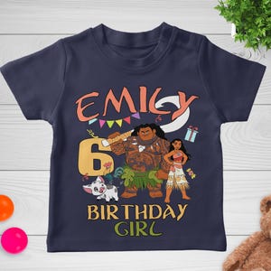 May include: A navy blue short-sleeve t-shirt featuring a graphic print with the name EMILY, the number 6, and the text BIRTHDAY GIRL. The design includes illustrations of two animated characters, a small pig, a gift box, and colorful bunting flags.