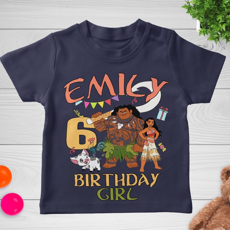 May include: A navy blue short-sleeve t-shirt featuring a graphic print with the name EMILY, the number 6, and the text BIRTHDAY GIRL. The design includes illustrations of two animated characters, a small pig, a gift box, and colorful bunting flags.