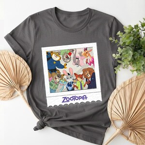 May include: Dark gray t-shirt featuring a Zootopia-themed graphic. The graphic is a polaroid-style image with various cartoon animal characters. The word "Zootopia" is printed in a purple font at the bottom of the image.