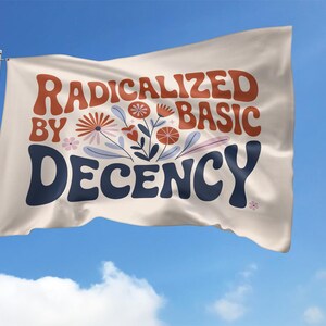 May include: A white flag with the words "RADICALIZED BY BASIC DECENCY" in orange and navy blue, with floral illustrations, waving against a blue sky. The flag is on a pole.