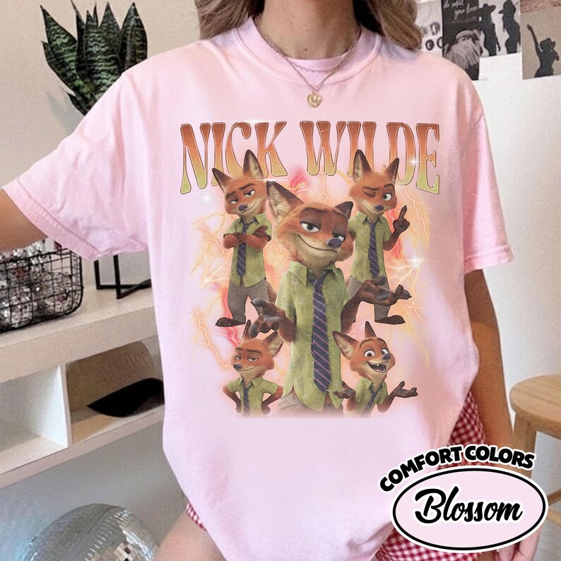 May include: A light pink crew neck t-shirt featuring a graphic print of the character Nick Wilde in various poses. The text NICK WILDE is printed in a curved, retro-style font above the character illustrations, which are set against a yellow lightning background.