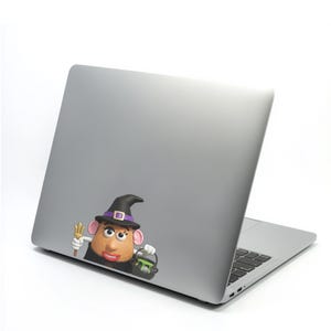 May include: A silver laptop with a colorful sticker of a potato-shaped character dressed as a witch. The character wears a black witch hat with a purple band, and holds a trident and a cauldron with green liquid.