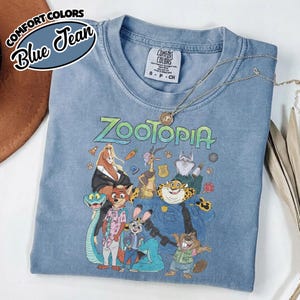 May include: A blue jean colored short-sleeve t-shirt featuring a graphic print of various Zootopia characters including Nick Wilde, Judy Hopps, and Chief Bogo. The shirt has a crew neckline and a Comfort Colors brand label on the inside collar.