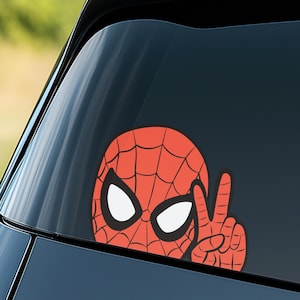 Pu&ograve; includere: Un adesivo con il volto di Spider-Man rosso e nero con forme di occhi bianchi e una mano che fa il segno della pace. L'adesivo &egrave; su un finestrino dell'auto.