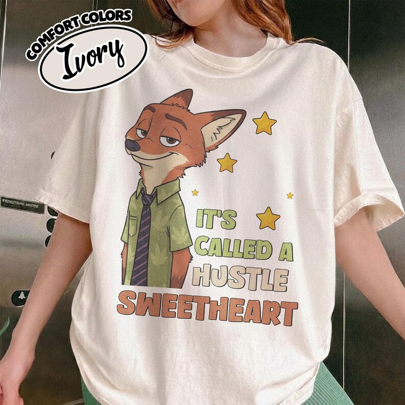 Puede incluir: Una camiseta de color marfil de manga corta con el gr&aacute;fico de un zorro de dibujos animados con camisa verde y corbata a rayas. El texto dice IT'S CALLED A HUSTLE SWEETHEART, acompa&ntilde;ado de estrellas amarillas. La camiseta tiene cuello redondo.