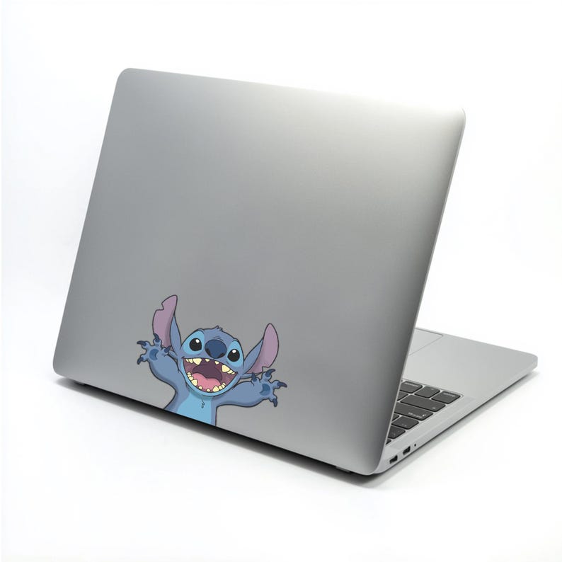 May include: A silver laptop with a cartoon image of Stitch, a blue alien character with large ears and a wide grin. The laptop is open, revealing a keyboard. The image is a sticker or decal applied to the laptop's exterior.