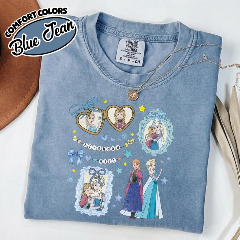 May include: A blue jean colored Comfort Colors t-shirt featuring a graphic print of two animated characters, Elsa and Anna. The design includes heart-shaped lockets, ornate frames, blue bows, stars, and the text "Birthday girl" on a string.