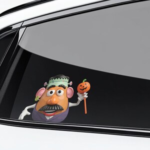 May include: A Halloween-themed Mr. Potato Head figure, dressed as Frankenstein, holding a small orange pumpkin on a stick. The figure has a black mustache, a purple vest, and a green and black hat. The background is a car window.