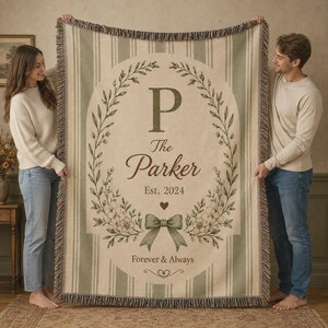 May include: A woven throw blanket featuring a cream and sage green striped pattern. The center displays a botanical wreath surrounding the letter P, the text The Parker, Est. 2024, a small heart, a bow illustration, and Forever & Always.