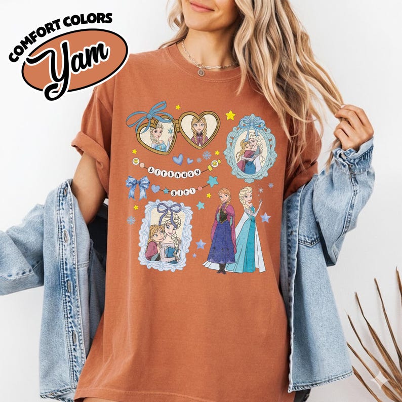 May include: A rust-colored t-shirt featuring a graphic print of characters from the movie Frozen, including heart-shaped lockets, ornate frames, and the text "birthday girl" on a string of beads, with a Comfort Colors Yam logo in the corner.