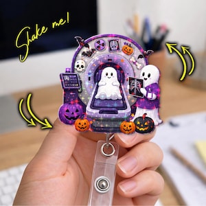 May include: A circular badge reel featuring a purple and black Halloween-themed design with a ghost inside an MRI machine, a ghost doctor holding an X-ray, pumpkins, a skull, and bats. The text "Shake me!" is written in yellow with yellow arrows.