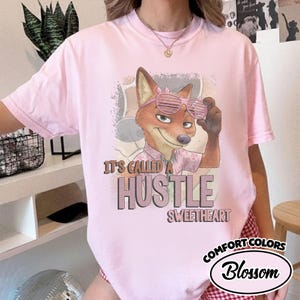 Puede incluir: Una camiseta rosa claro con un gr&aacute;fico de un zorro de dibujos animados que lleva gafas de sol de persiana rosas y una camisa con cuello. El texto dice "IT'S CALLED A HUSTLE SWEETHEART" en una fuente marr&oacute;n negrita. Tiene cuello redondo.