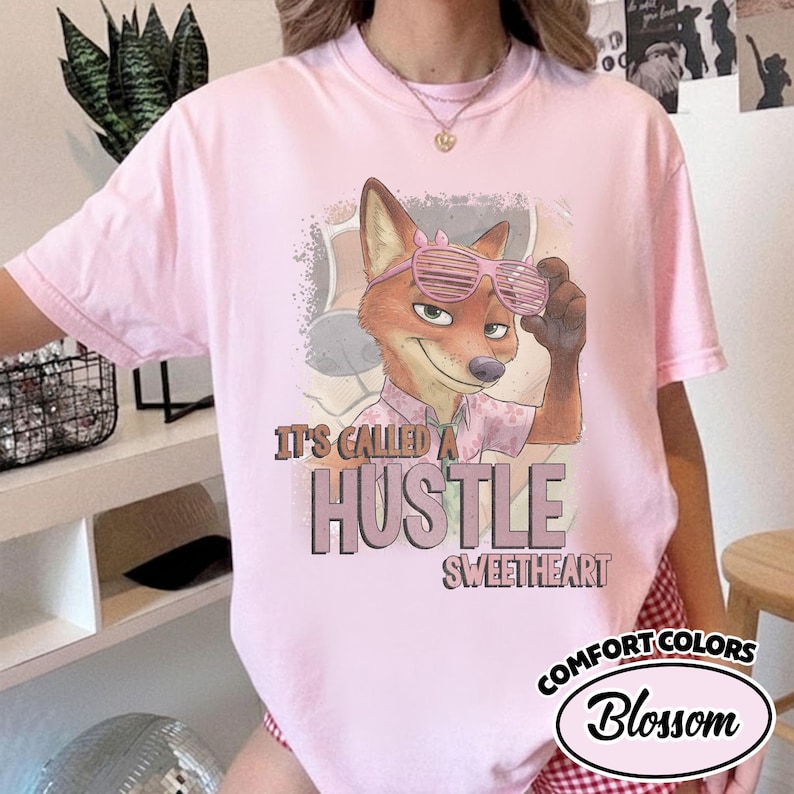 Puede incluir: Una camiseta rosa claro con un gr&aacute;fico de un zorro de dibujos animados que lleva gafas de sol de persiana rosas y una camisa con cuello. El texto dice "IT'S CALLED A HUSTLE SWEETHEART" en una fuente marr&oacute;n negrita. Tiene cuello redondo.
