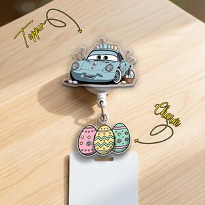 May include: A badge reel with a light blue cartoon car topper and three Easter egg charms. The car has a basket of eggs and a colorful egg on top. The charms are pink, yellow, and blue with floral and geometric designs. The reel has a white card holder.
