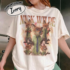 May include: An ivory t-shirt featuring a graphic print of the character Nick Wilde from Zootopia in various poses. The text NICK WILDE appears in a large, arched, distressed font above the character illustrations. A Comfort Colors Ivory label is visible.