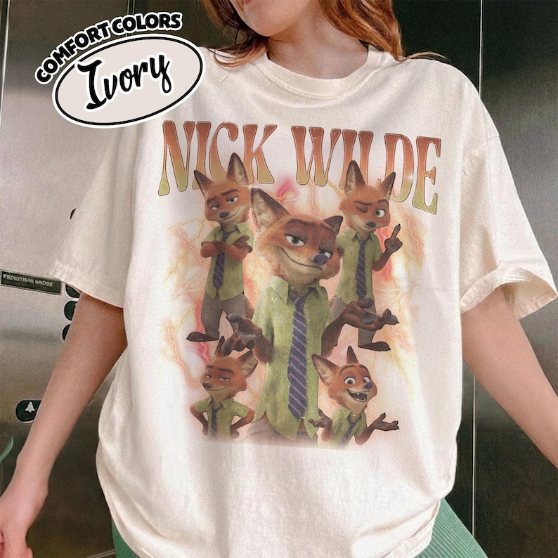 May include: An ivory t-shirt featuring a graphic print of the character Nick Wilde from Zootopia in various poses. The text NICK WILDE appears in a large, arched, distressed font above the character illustrations. A Comfort Colors Ivory label is visible.