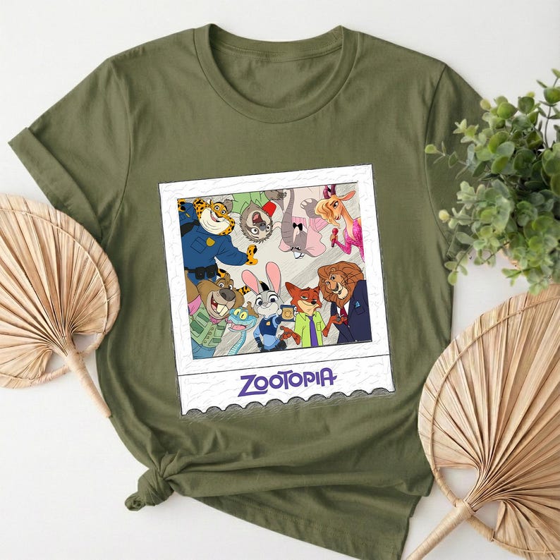 May include: Olive green t-shirt featuring a graphic of characters from the animated film Zootopia. The graphic is in a white frame with the word "Zootopia" in blue text at the bottom. The shirt is made of a soft material.