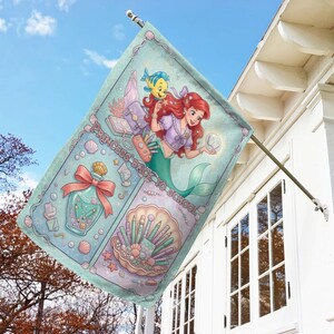 Puede incluir: Bandera decorativa para exteriores con una ilustraci&oacute;n de sirena de pelo rojo y un pez amarillo. La bandera tiene un fondo azul claro con el texto "PRINCESS BACK TO SCHOOL". Tambi&eacute;n presenta un frasco de perfume y brochas de maquillaje.