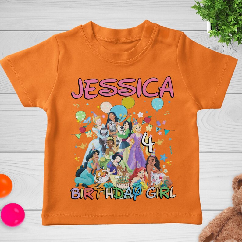 May include: Orange short-sleeved t-shirt with the name "JESSICA" in pink letters. The shirt features a colorful graphic of Disney princesses, the number "4", and the words "BIRTHDAY GIRL".