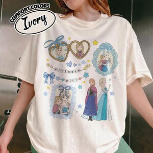 May include: An ivory short-sleeve t-shirt featuring a graphic print of two cartoon characters in heart-shaped and rectangular frames, a banner with the text "birthday girl," blue bows, stars, and snowflakes. The shirt is labeled Comfort Colors Ivory.