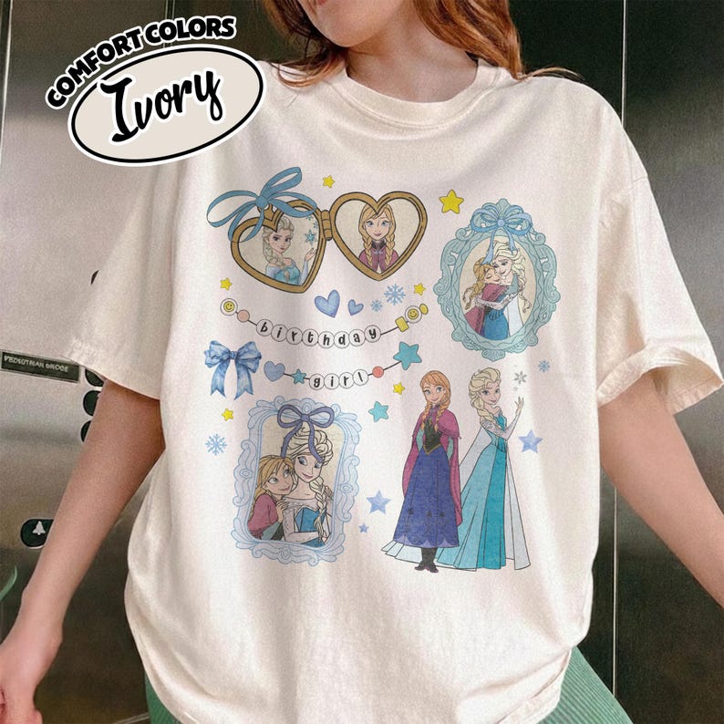 May include: An ivory short-sleeve t-shirt featuring a graphic print of two cartoon characters in heart-shaped and rectangular frames, a banner with the text "birthday girl," blue bows, stars, and snowflakes. The shirt is labeled Comfort Colors Ivory.