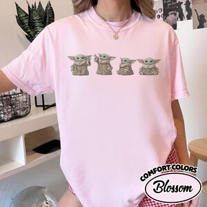 Puede incluir: Una camiseta de manga corta de color rosa claro con un estampado gr&aacute;fico de cuatro peque&ntilde;as criaturas verdes de orejas grandes con t&uacute;nicas de color tostado en varias poses. La camiseta tiene cuello redondo y el texto COMFORT COLORS Blossom.