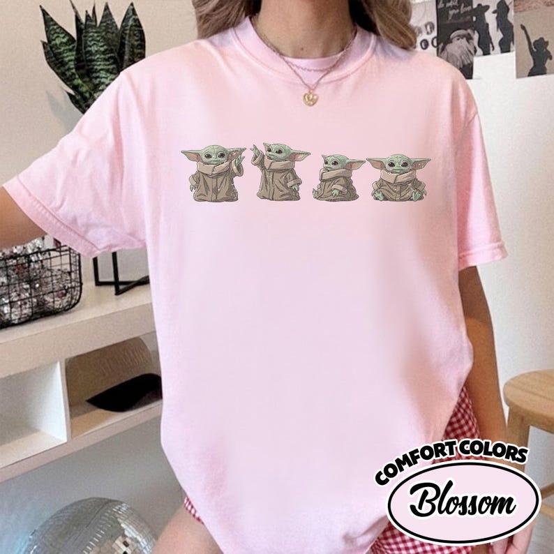Puede incluir: Una camiseta de manga corta de color rosa claro con un estampado gr&aacute;fico de cuatro peque&ntilde;as criaturas verdes de orejas grandes con t&uacute;nicas de color tostado en varias poses. La camiseta tiene cuello redondo y el texto COMFORT COLORS Blossom.