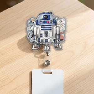 May include: A clear badge reel featuring a detailed illustration of the R2-D2 droid from Star Wars. The design includes blue, white, and silver colors, with a clear plastic reel and a white rectangular tag.