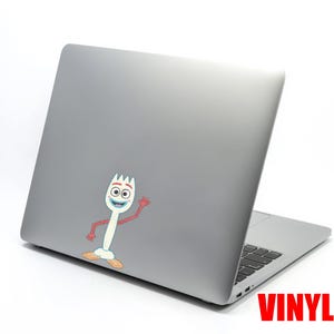 May include: A gray laptop with a colorful vinyl sticker of the character Forky from Toy Story. The sticker is white with a fork head, blue mouth, and red arms. The word "VINYL" is printed in red.