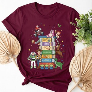 Może przedstawiać: Bordowa koszulka z kolorową grafiką postaci Disneya. Projekt przedstawia stos książek z tytułami takimi jak "Toy Story" i "Kopciuszek". Postacie to m.in. Woody, Buzz Lightyear i mali zieloni ludziki.