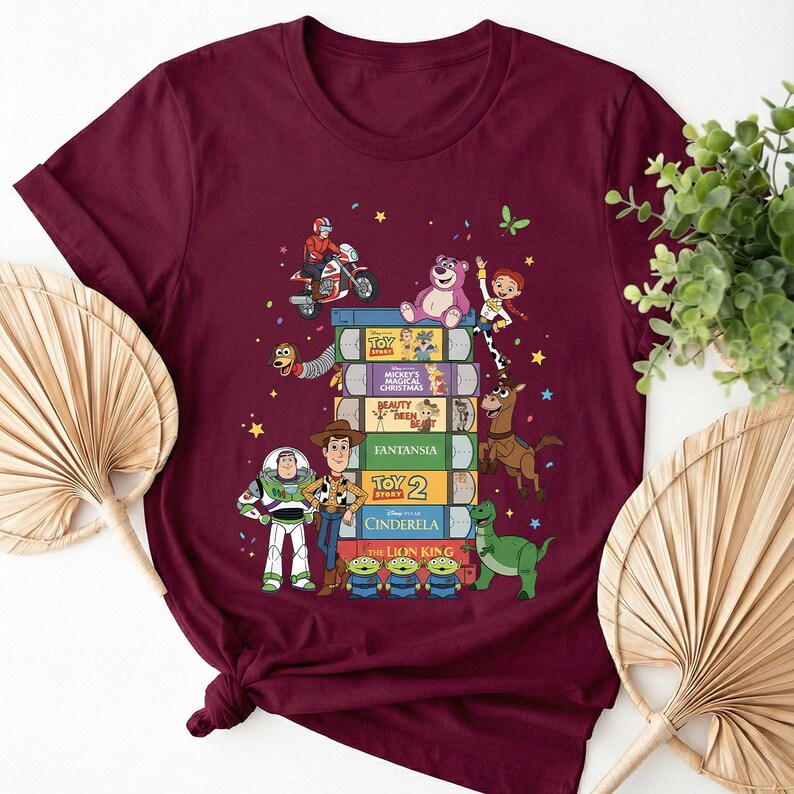 Może przedstawiać: Bordowa koszulka z kolorową grafiką postaci Disneya. Projekt przedstawia stos książek z tytułami takimi jak "Toy Story" i "Kopciuszek". Postacie to m.in. Woody, Buzz Lightyear i mali zieloni ludziki.