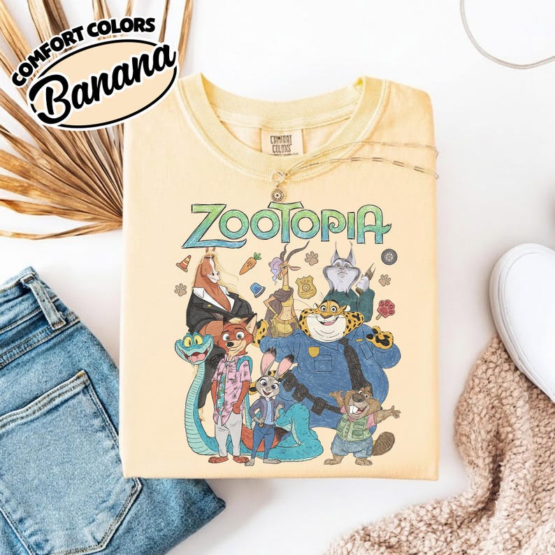 May include: A pale yellow t-shirt featuring a graphic print of various Zootopia characters including Nick Wilde, Judy Hopps, and Chief Bogo. The text Zootopia is printed in a stylized green font above the characters, with small icons like a carrot and badge.