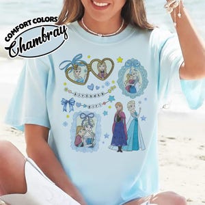 May include: A light blue t-shirt featuring a graphic print of characters from Frozen, including Anna and Elsa in various frames, a heart-shaped locket, and the text "birthday girl" on a string of beads. The shirt has a Comfort Colors Chambray label.