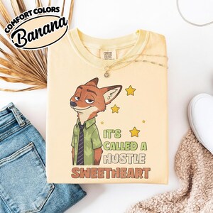 Puede incluir: Una camiseta de color amarillo p&aacute;lido con el gr&aacute;fico de un zorro de dibujos animados que lleva una camisa verde y una corbata oscura. El texto dice "IT'S CALLED A HUSTLE SWEETHEART", junto a estrellas amarillas. La camiseta tiene cuello redondo.