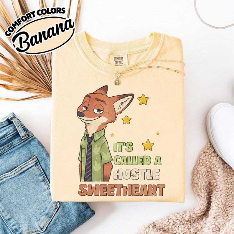 Puede incluir: Una camiseta de color amarillo p&aacute;lido con el gr&aacute;fico de un zorro de dibujos animados que lleva una camisa verde y una corbata oscura. El texto dice "IT'S CALLED A HUSTLE SWEETHEART", junto a estrellas amarillas. La camiseta tiene cuello redondo.