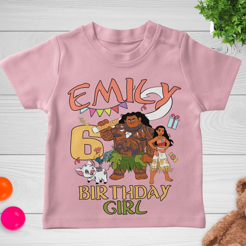 May include: A light pink short-sleeve t-shirt featuring a graphic print of Moana characters, a large number 6, and the text EMILY BIRTHDAY GIRL. The design includes a pig, a rooster, a hook, colorful bunting, and a gift box in a cartoon style.