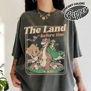 May include: A charcoal gray short-sleeve t-shirt featuring a graphic print of characters from The Land Before Time. The design includes a central long-necked dinosaur, a triceratops, a pterodactyl, and a small green dinosaur within a rectangular frame.