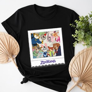 May include: Black t-shirt featuring a colorful graphic of characters from the animated movie "Zootopia" inside a polaroid frame. The word "Zootopia" is printed in purple script below the image. The shirt is made of a soft material.