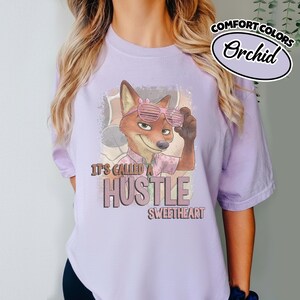 Puede incluir: Una camiseta de color morado claro con el gr&aacute;fico de un zorro de dibujos animados con gafas de sol rosas y una camisa estampada. El texto dice "IT'S CALLED A HUSTLE SWEETHEART" en letra negrita. Una etiqueta ovalada indica "COMFORT COLORS Orchid".