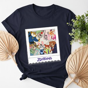 May include: Navy blue t-shirt featuring a graphic of characters from the animated film "Zootopia" in a polaroid-style frame. The word "Zootopia" is printed in a stylized font below the image. The shirt is made of a soft material.