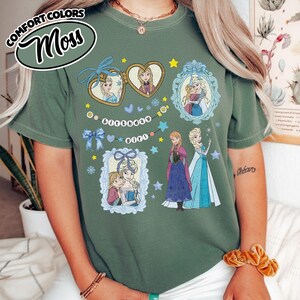 May include: A moss green t-shirt featuring a graphic print of two animated characters in various frames, including heart-shaped and ornate blue borders. The design includes the text "birthday girl" on a string, along with stars and snowflakes.