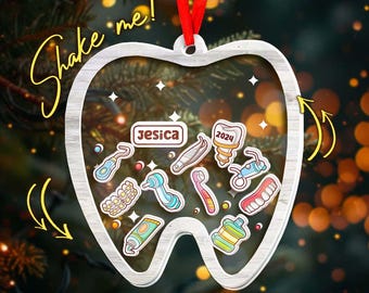 May include: A silver-colored, tooth-shaped Christmas ornament with a red ribbon. Inside the tooth shape are various dental-themed stickers, the name "Jesica", and the year "2024". The words "Shake me!" are written in a yellow, cursive font.