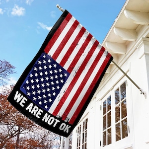 May include: A black flag with a distressed American flag design and the text "We Are Not Ok" in white lettering.