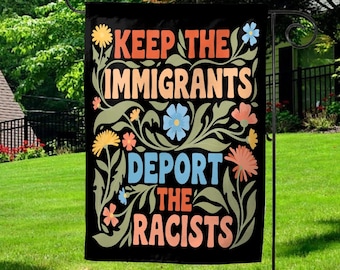 May include: Black garden flag with the text "KEEP THE IMMIGRANTS, DEPORT THE RACISTS" in colorful, stylized lettering. The text is surrounded by floral illustrations in shades of green, yellow, orange, and blue. The flag is hanging on a black metal stand.