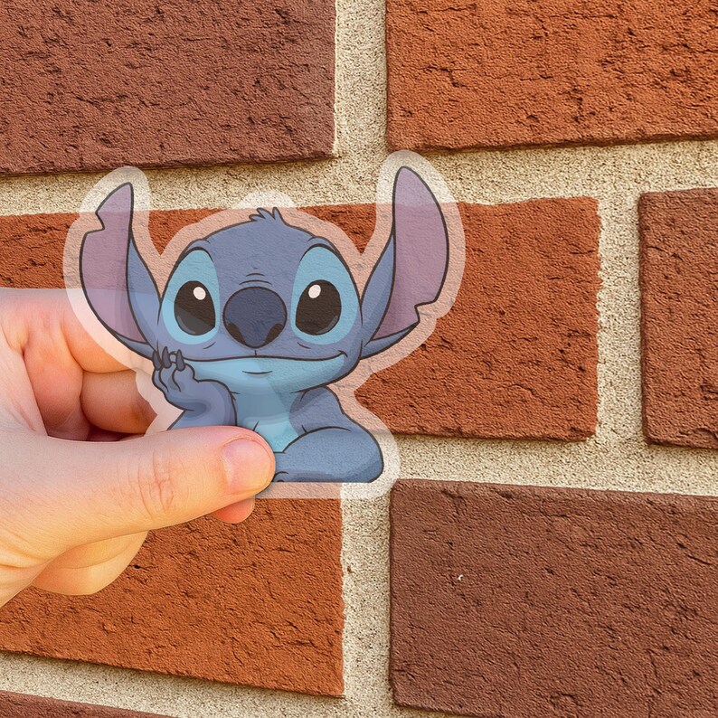 May include: A transparent sticker featuring a blue cartoon character with large ears and big eyes. The character is resting its chin on its hand. The sticker is held in front of a brick wall.