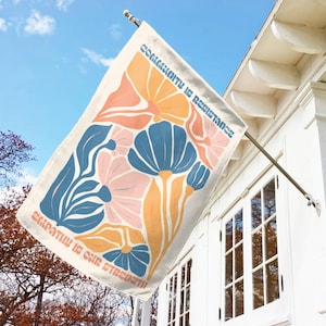 May include: A rectangular outdoor flag with a floral design in shades of blue, peach, and yellow. The flag reads "Community is Resistance" and "Empathy is Our Strength" in a retro font. The flag is mounted on a white house.